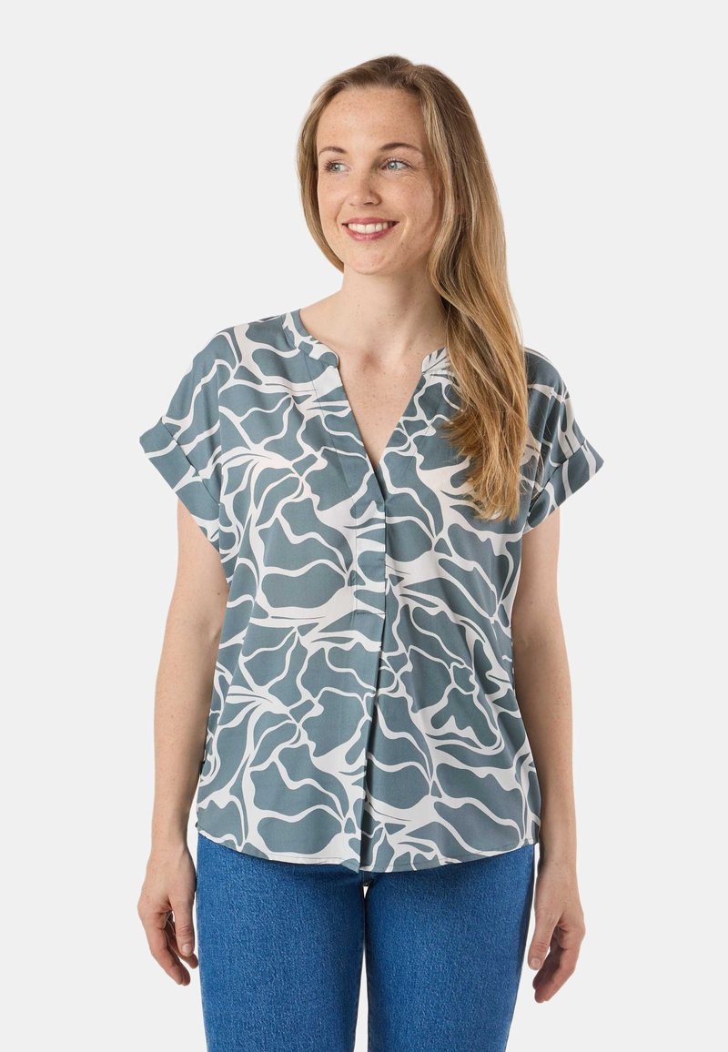 Femme aux longs cheveux blonds portant un chemisier à manches courtes bleu et blanc à motifs, et un jean bleu, souriante et regardant légèrement sur le côté.