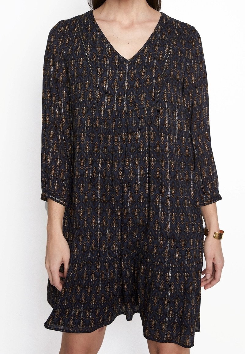 Femme portant une robe bleu foncé à motifs, manches longues et col en V, accessoirisée d'un bracelet en or au poignet droit.