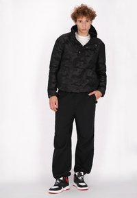 Schwarze Camouflage-Daunenjacke mit hohem Kragen, kombiniert mit lockeren schwarzen Hosen und schwarzen Turnschuhen mit roten Akzenten.