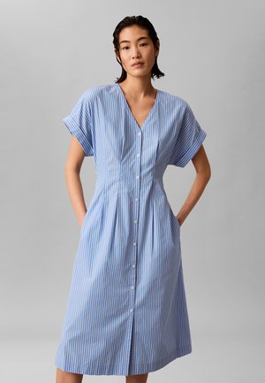 Jonge vrouw staat met handen in zakken, draagt een blauwe en witte midi jurk met verticale strepen, knopen en korte mouwen.