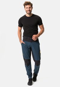 Pantalons de plein air pour hommes en bleu avec des accents noirs, fabriqués en tissu léger, dotés de poches zippées et de genoux renforcés. Chemise noire portée.
