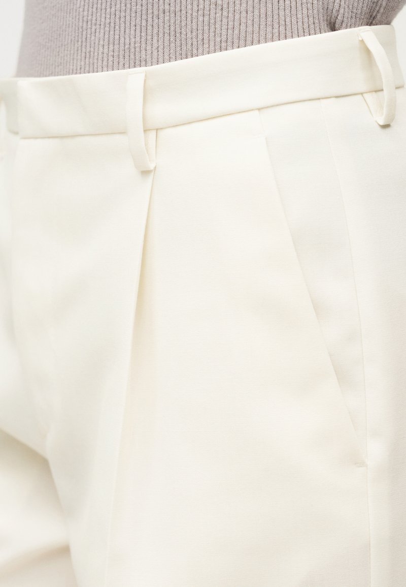 Pantalon de couleur crème avec devant plat, plis simples et passants pour ceinture. Le tissu a une texture lisse avec une coupe ajustée.