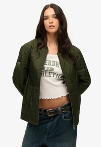 Gequiltede olijfgroene bomberjack met een rits aan de voorkant, gedragen over een witte cropped top. Het model combineert het met high-waisted blauwe jeans en een gesp met studs.
