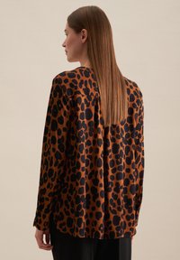 Camicia a maniche lunghe in tessuto marrone con stampa leopardata nera. Presenta una linea dritta e un delicato dettaglio plissettato sul retro.