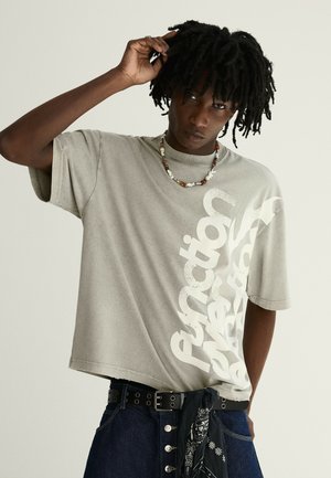 Ung mand med mørke dreadlocks iført en beige oversized t-shirt med hvid tekst, en perlehalskæde, mørke jeans og et sort bandana-bælte.