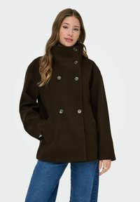 Abrigo marrón de doble botonadura con grandes botones, cuello alto, bolsillos laterales y un corte relajado, confeccionado en un material suave y grueso.