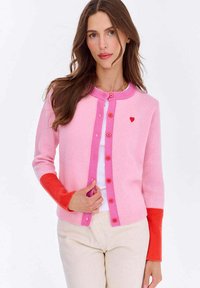 Dzianinowy kardigan w kolorze różowym i koralowym, z okrągłym dekoltem, zapięciem na guziki oraz małym czerwonym serduszkiem na klatce piersiowej. Gładka struktura.