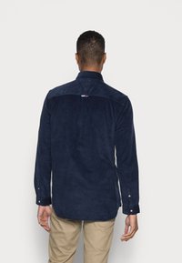 Tmavě modrá manšestrová košile s špičatým límečkem, knoflíkovými manžetami a malým víceměrným logem na zádech. Měkká textura a střih na míru.
