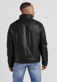 Schwarze Lederjacke mit hohem Kragen, taillierter Passform und genähten Details. Jede Ärmel hat ein leicht gepolstertes Design. Getragen mit hellblauen Jeans.