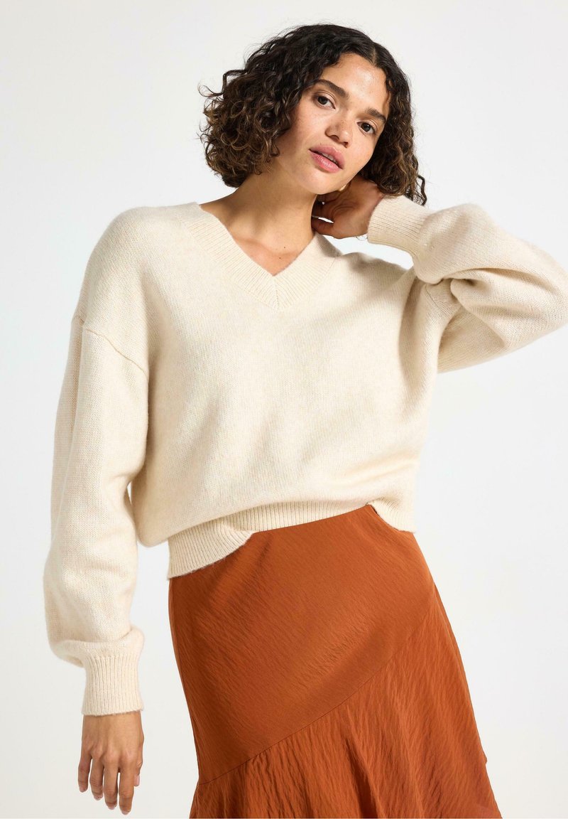 Una donna con i capelli ricci indossa un maglione crema oversize e una gonna color ruggine, posando con una mano dietro il collo su uno sfondo neutro.
