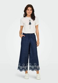 Brede donkerblauwe broeken met witte bloemembroideries aan de onderkant, gecombineerd met een witte t-shirt met een grafisch ontwerp.