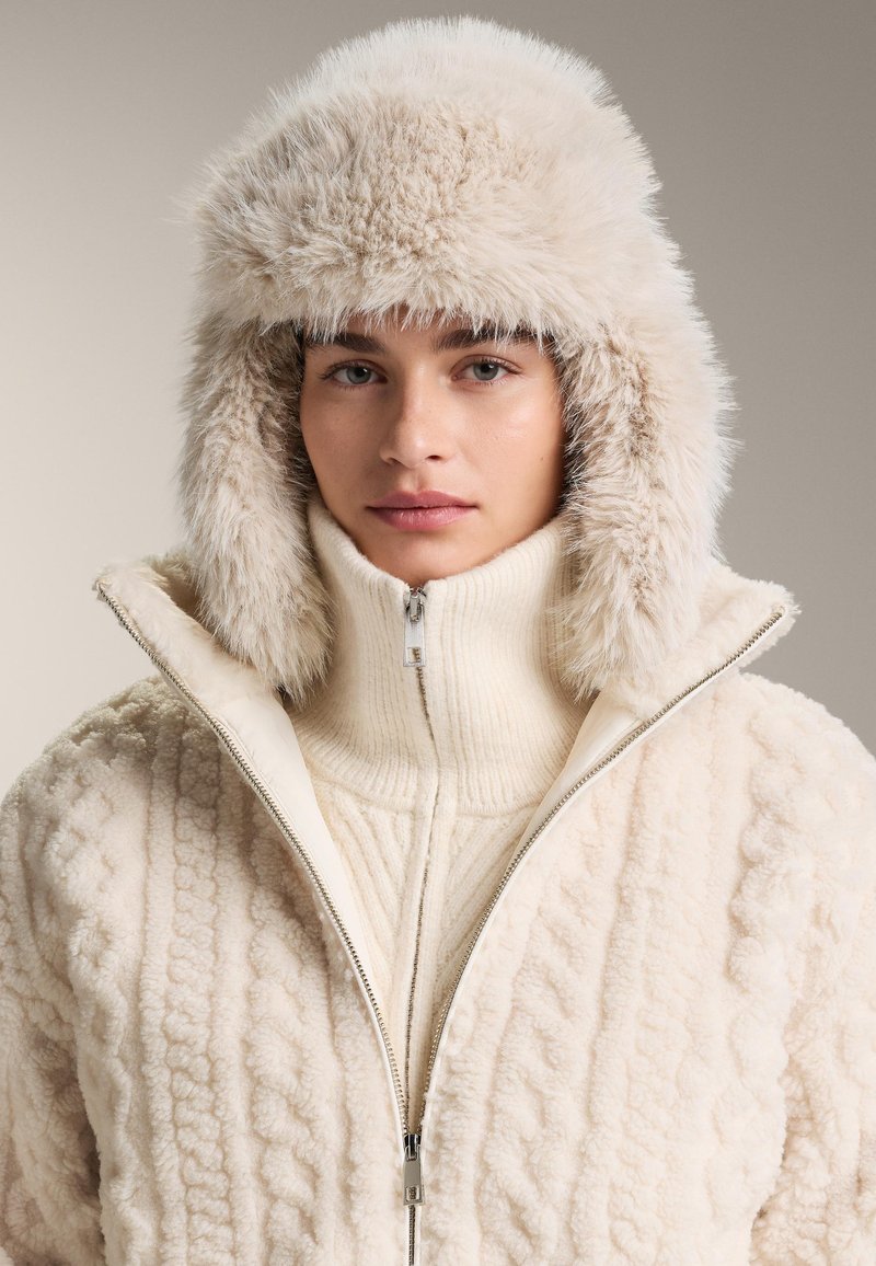Žena v krémovej chlpaté trapper čiapke a textúrovanej bunde na zips, hľadiaca priamo do kamery s neutrálnym výrazom.