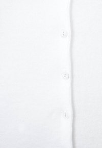 Vit kofta i stickad tyg, med en knäppt framtill som har fem vita knappar. Slät textur och enkel design utan mönster.