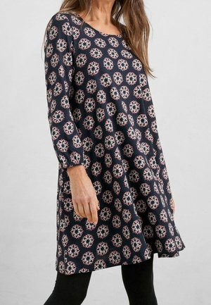 Woman wearing navy blue long-sleeve dress with pink and white floral circular patterns, paired with black leggings, standing against plain background.