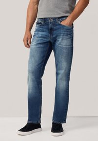 Hombre vistiendo jeans desgastados de color azul, camiseta gris, zapatillas negras con suelas blancas, de pie con una mano en el bolsillo contra un fondo sencillo.