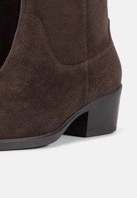 Gros plan d'une botte cheville en daim marron avec un talon bas empilé et des détails de couture visibles.
