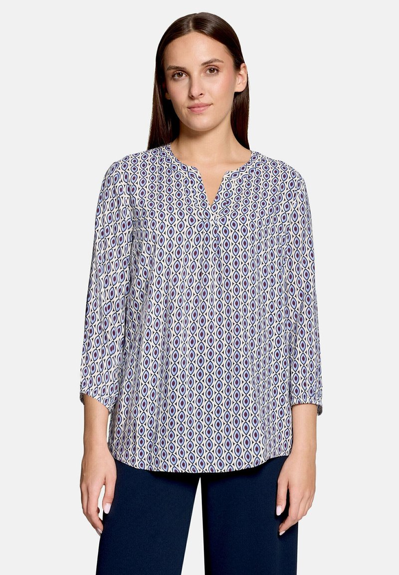 Femme portant un chemisier blanc à manches longues avec un motif géométrique bleu et violet et un pantalon bleu foncé, debout devant un fond uni.