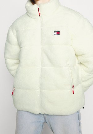 Persoon draagt een wit fleece gewatteerd jack met rode ritsen en een Tommy Jeans-logo op de borst, gecombineerd met lichtblauwe spijkerbroek.