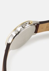 Orologio di tonalità argento con cassa rotonda, dettagli dorati e cinturino in pelle marrone. Presenta due pulsanti e una finitura metallica liscia.