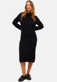 Femme avec de longs cheveux portant une robe midi noire à col roulé et des mocassins noirs, se tenant les mains derrière le dos sur un fond blanc.