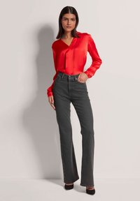 Rote Satinbluse mit V-Ausschnitt, kombiniert mit hochgeschnittenen, ausgestellten dunkelblauen Jeans. Das Model steht in schwarzen High Heels vor einem neutralen Hintergrund.