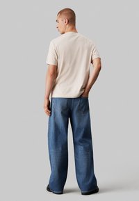Camiseta de color beige claro con un corte holgado, combinada con jeans de mezclilla de pernera ancha y lavado medio que cuentan con bolsillos traseros y detalles de costura mínimos.