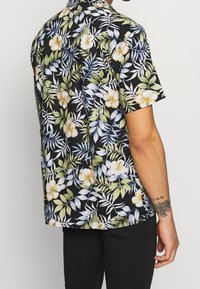 Korte mouw overhemd in zwarte stof met een bloemenpatroon van witte en gele bloemen en groene bladeren, ontspannen pasvorm met een kraag.