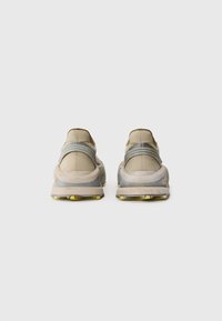 Baskets de sport beiges avec une tige texturée, des accents gris et une semelle en caoutchouc avec un motif jaune. Design minimaliste avec une forme arrondie.