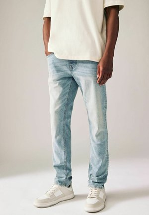 Vaqueros de mezclilla azul claro con un ajuste relajado, que presentan un desgaste sutil y arrugas. Combinados con zapatillas blancas y una blusa de manga corta color crema.