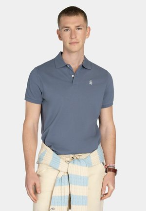 Joven con una camisa tipo polo azul y pantalones beige, con un suéter azul y blanco a rayas atado alrededor de la cintura, posando frente a un fondo blanco.