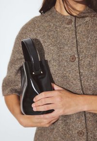 Melnas lakotas ādas rokassomi ar izliekto rokturi, novietota uz tekstūrēta brūna adīta džempera ar pogām. Kārtīga, strukturēta forma.