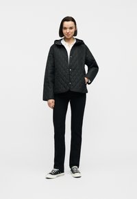 Νεαρή γυναίκα που φοράει μαύρο καπιτονέ μπουφάν, λευκό πουκάμισο, μαύρο παντελόνι και μαύρα αθλητικά παπούτσια, στέκεται με το ένα χέρι στην τσέπη σε λευκό φόντο.