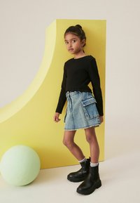 Svart långärmad topp, blå denimkjol med fickor, svarta ankelstövlar, grå strumpor, med en grön boll och gul bakgrund.