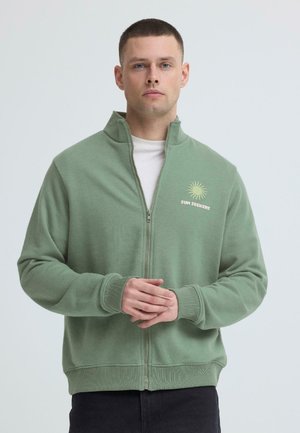 Muškarac s kratkom kosom u zelenoj flis majici na patentni zatvarač s natpisom "Sun Seekers" i slikom sunca, stoji ispred jednobojne svijetle pozadine.