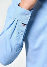 Világoskék, hosszú ujjú ing, gombos mandzsettával, pliszírozással és egy kis hímzett részlettel piros, fehér és fekete színben az ujján.