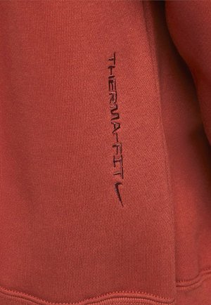 Tejido Therma-FIT rojo con textura acanalada. Presenta un destacado logo y texto bordados en negro, con un diseño ajustado y sin costuras.