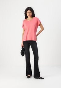 Top roz cu mâneci scurte și croială lejeră, asortat cu pantaloni negri evazați și pantofi negri fără toc. Modelul ține o geantă neagră.