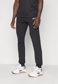 Schwarze tapered Sweatpants mit weicher Textur, ausgestattet mit Seitentaschen und einem gestickten Logo am oberen Oberschenkel. Getragen mit weißen Sportschuhen.