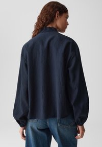 Camicia blu scuro con finitura testurizzata, colletto alto e maniche lunghe a sbuffo, abbinata a jeans blu.