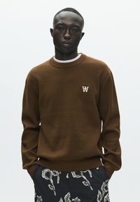 Pull en maille marron avec un col rond et un logo "W" blanc sur la poitrine, associé à un short foncé à motifs. Texture lisse, design décontracté.