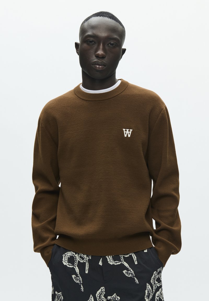 Pull en maille marron avec un col rond et un logo "W" blanc sur la poitrine, associé à un short foncé à motifs. Texture lisse, design décontracté.