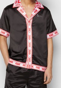 Person wearing a black satin short-sleeve shirt and shorts set with red and white laurel patterned trim and white buttons.