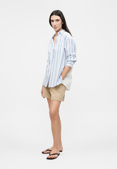 Γυναίκα που φοράει μπλε και λευκό ριγέ μπλουζάκι μακρυμάνικο, μπεζ σορτς και μαύρα σαγιονάρες, στέκεται με το ένα χέρι στην τσέπη.