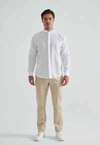 Chemise blanche à manches longues avec un col montant, associée à un pantalon beige et des baskets blanches, présentant une coupe ajustée et une texture lisse.