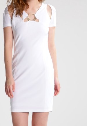 Femme portant une courte robe blanche avec des découpes aux épaules et un détail décoratif d'anneau métallique au niveau de l'encolure.
