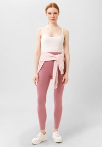 Witte tanktop, roze high-waisted leggings en een lichtroze sweater geknoopt in de taille. Witte sneakers met een gestructureerde zool. Neutrale achtergrond.