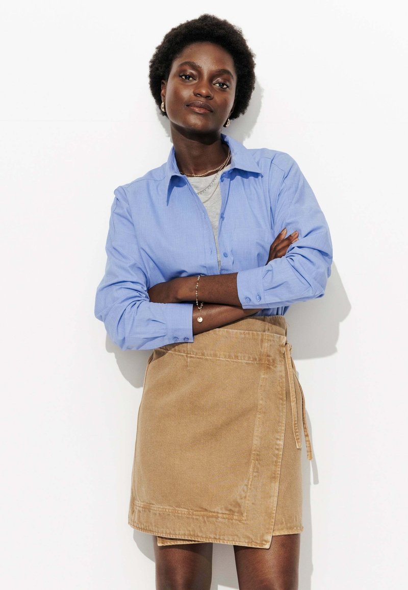 Chemise bleu clair à boutons portée avec une jupe portefeuille beige. Le tissu semble lisse avec des détails de couture visibles. Modèle se tenant avec les bras croisés.