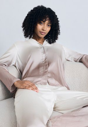 Γυναίκα με σγουρά μαλλιά που κάθεται σε έναν ανοιχτόχρωμο καναπέ, φορώντας μια χαλαρή, γυαλιστερή μπεζ μπλούζα και αντίστοιχο παντελόνι, κοιτάζοντας κατευθείαν προς την κάμερα.