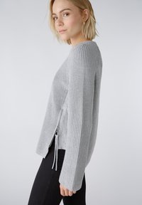 Pull en tricot gris clair avec une texture côtelée et un détail de fermeture éclair sur le côté. Il possède des manches longues et une coupe décontractée.