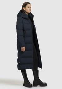 Lungo cappotto imbottito blu navy con cappuccio, caratterizzato da un design imbottito, cerniere laterali e una texture liscia, abbinato a stivali da combattimento neri.